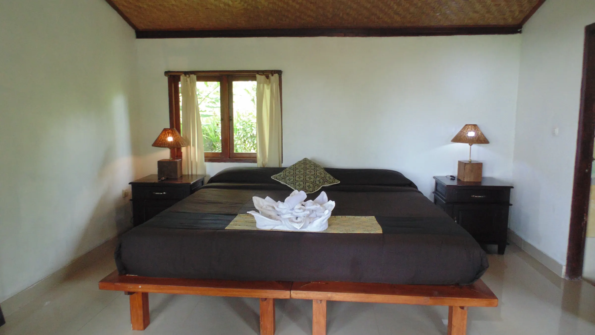Teak platform bed with grey linen and rattan lamps under a woven bamboo ceiling