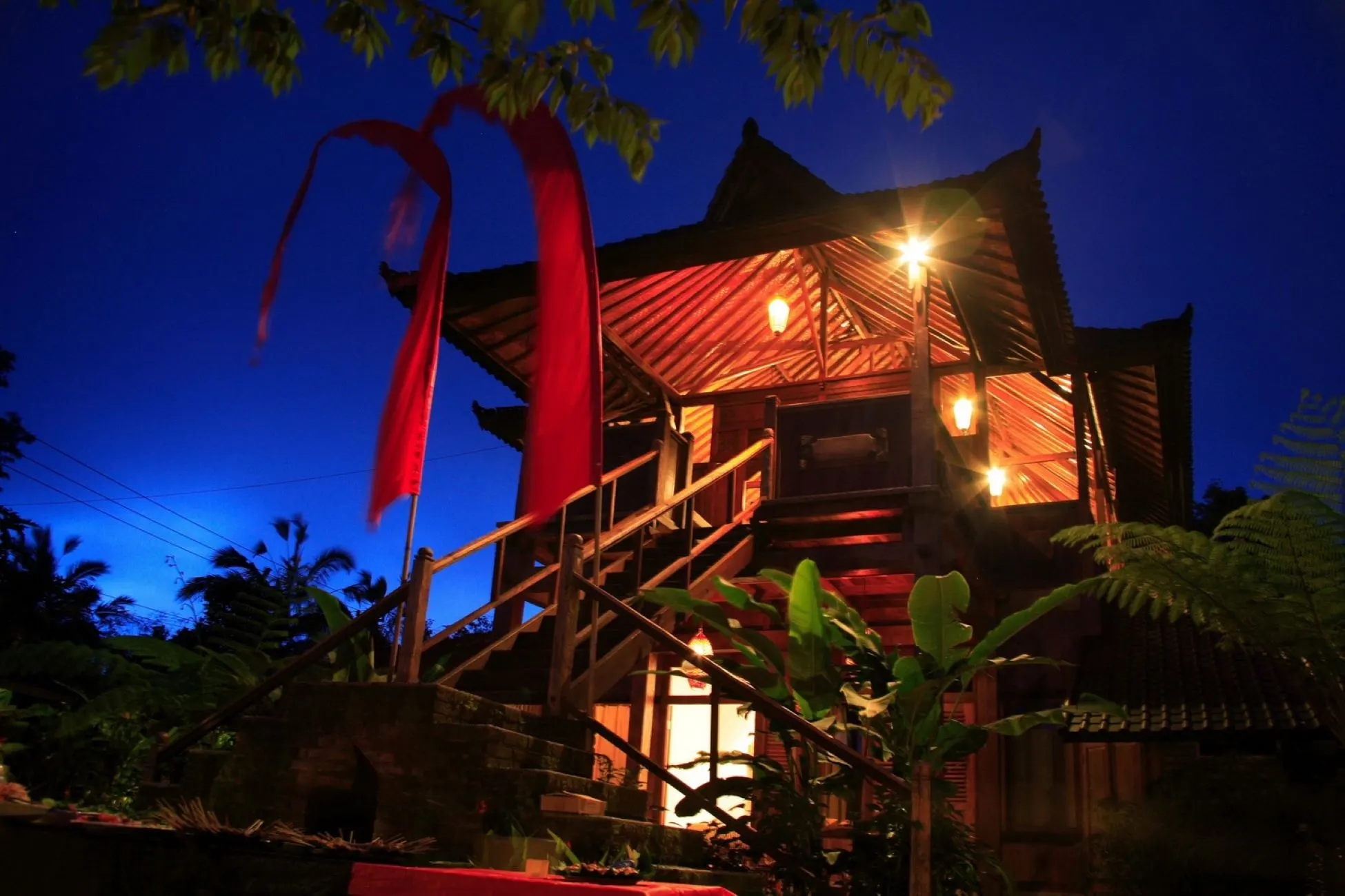 A timber house lit up at dusk, with red ceremonial flags against the blue sky