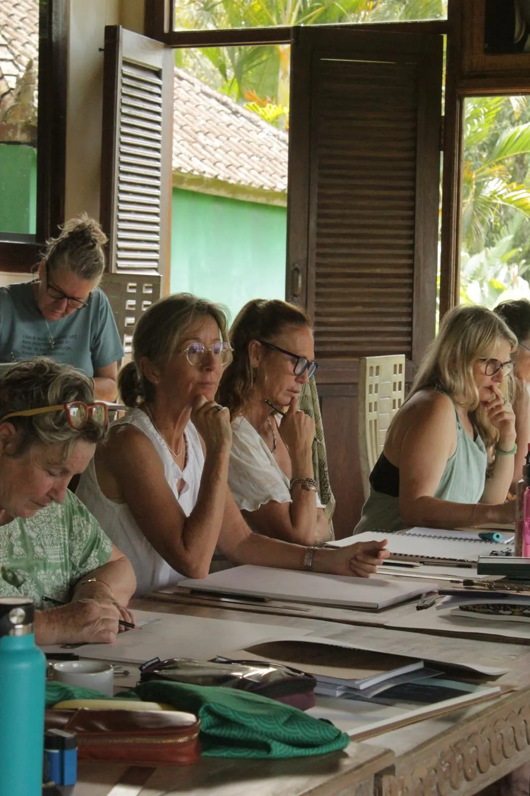 Guests listening along a carved wooden table, louvred teak shutters open behind