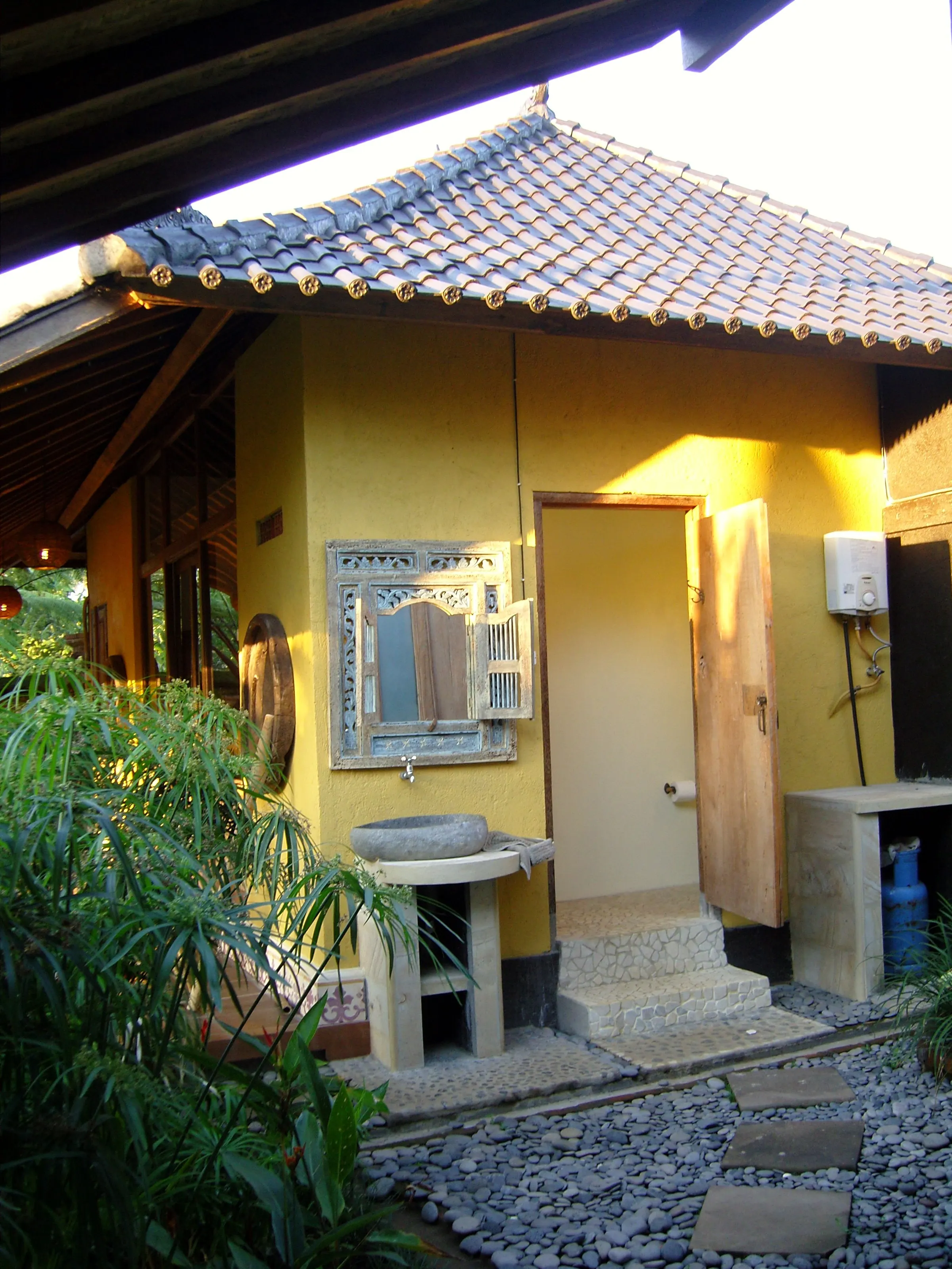 An open-air bathroom with a yellow wall, carved mirror and stone basin on a pebble path